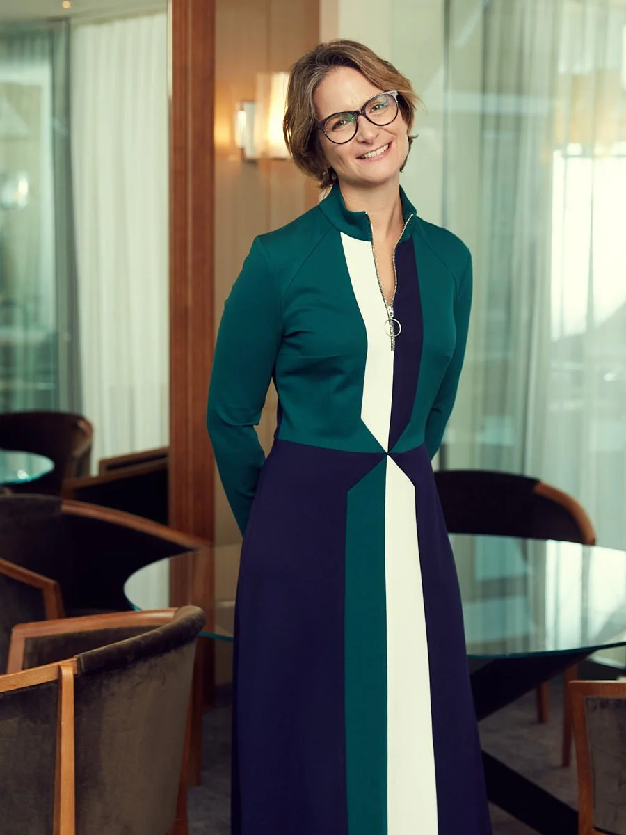 A woman with short brown hair and glasses, smiling, wearing a color-blocked teal, navy, and white dress, standing in a modern indoor space with wooden and glass elements.