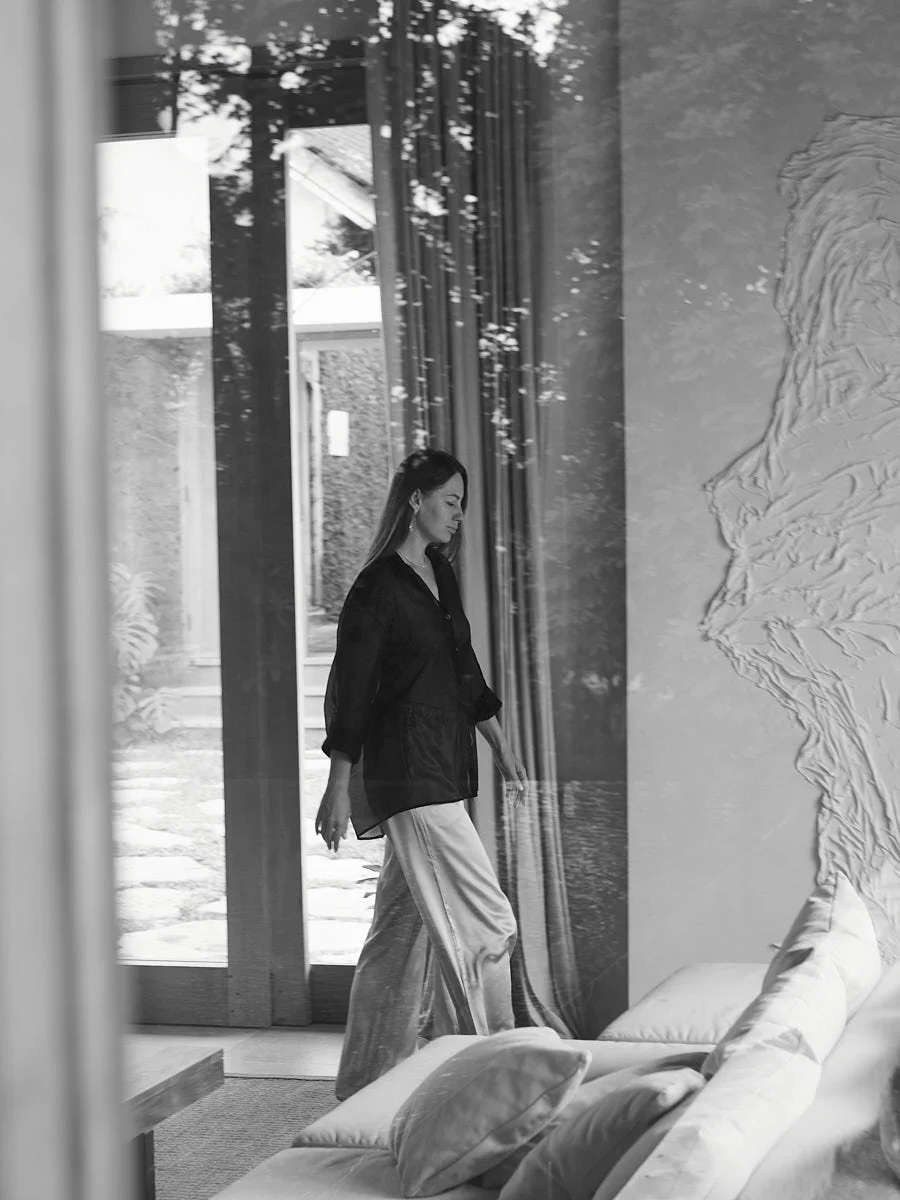 A woman with long hair, dressed in a dark top and light-colored pants, walking inside a modern home near a large glass door with curtains. Reflections of trees and a garden are visible outside.