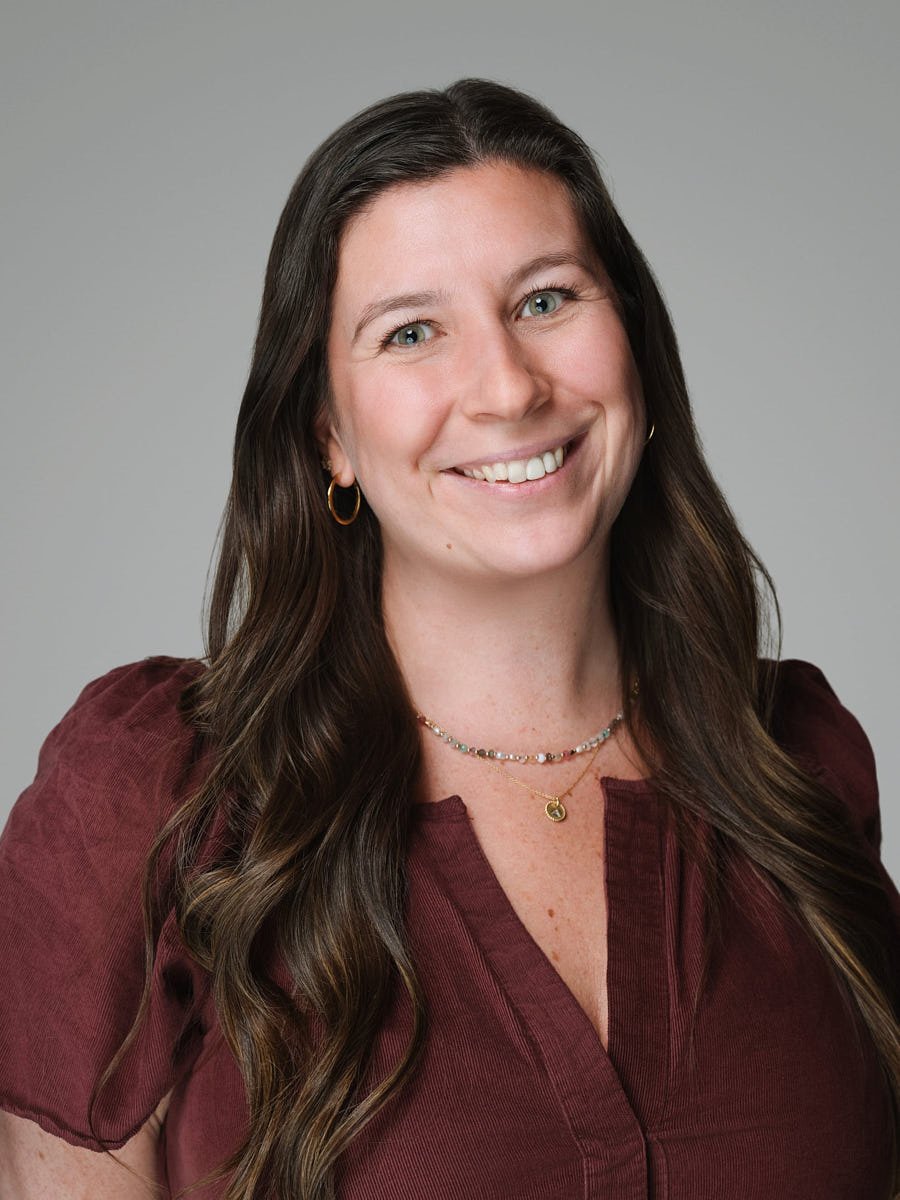 Portrait of a smiling woman with long brown hair, wearing a burgundy top, gold hoop earrings, a beaded necklace, and a gold pendant necklace, against a gray background by Match Production.
