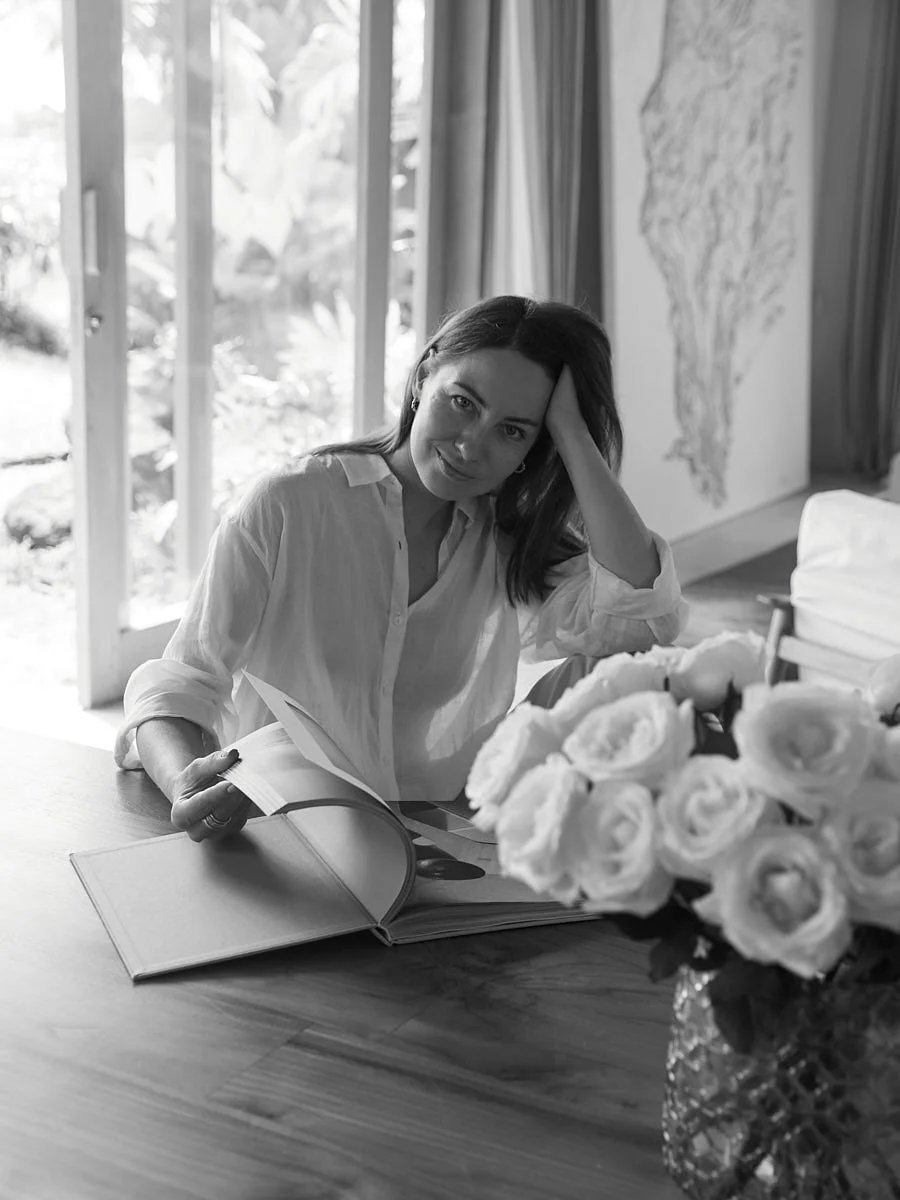 A woman sitting at a table with a large bouquet of roses, looking at a photo album, inside a room with large windows and curtains, black and white photo.