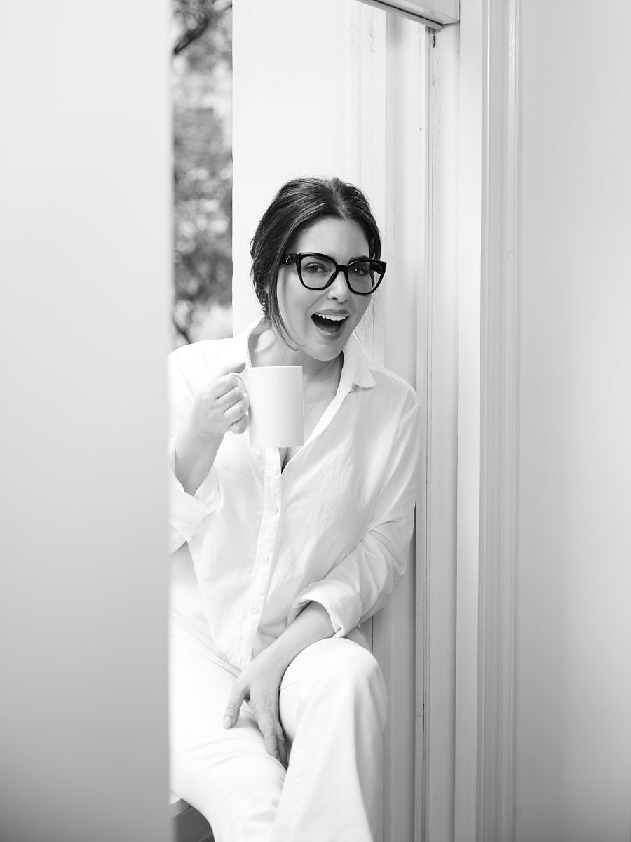 A woman with short dark hair and glasses, dressed in a white shirt, sitting by a window and holding a coffee mug, smiling and looking at the camera.
