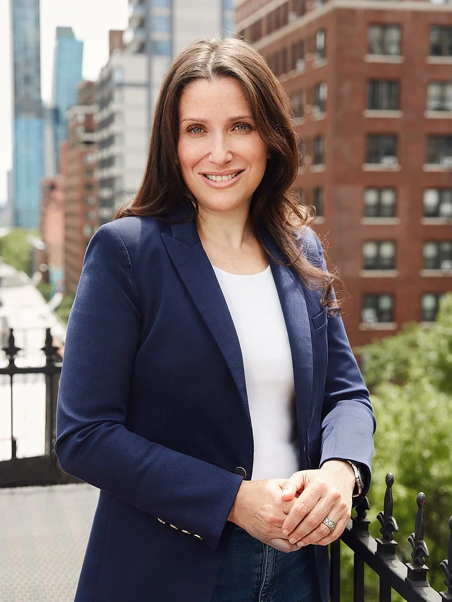 A woman with long brown hair smiling outdoors on a balcony in an urban setting. She is wearing a navy blue blazer over a white top.