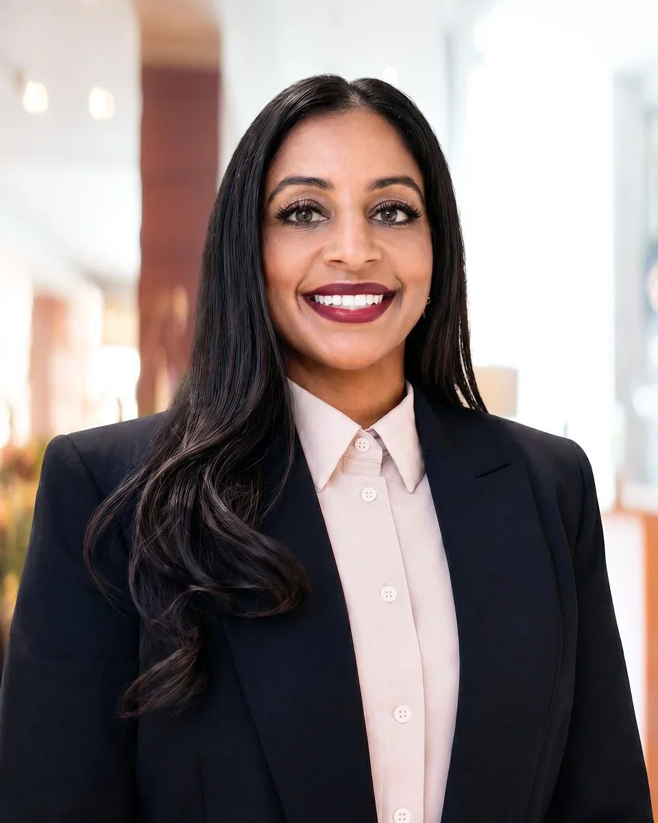 A professional woman with long dark hair, wearing a white blouse and a dark blazer, smiling indoors.