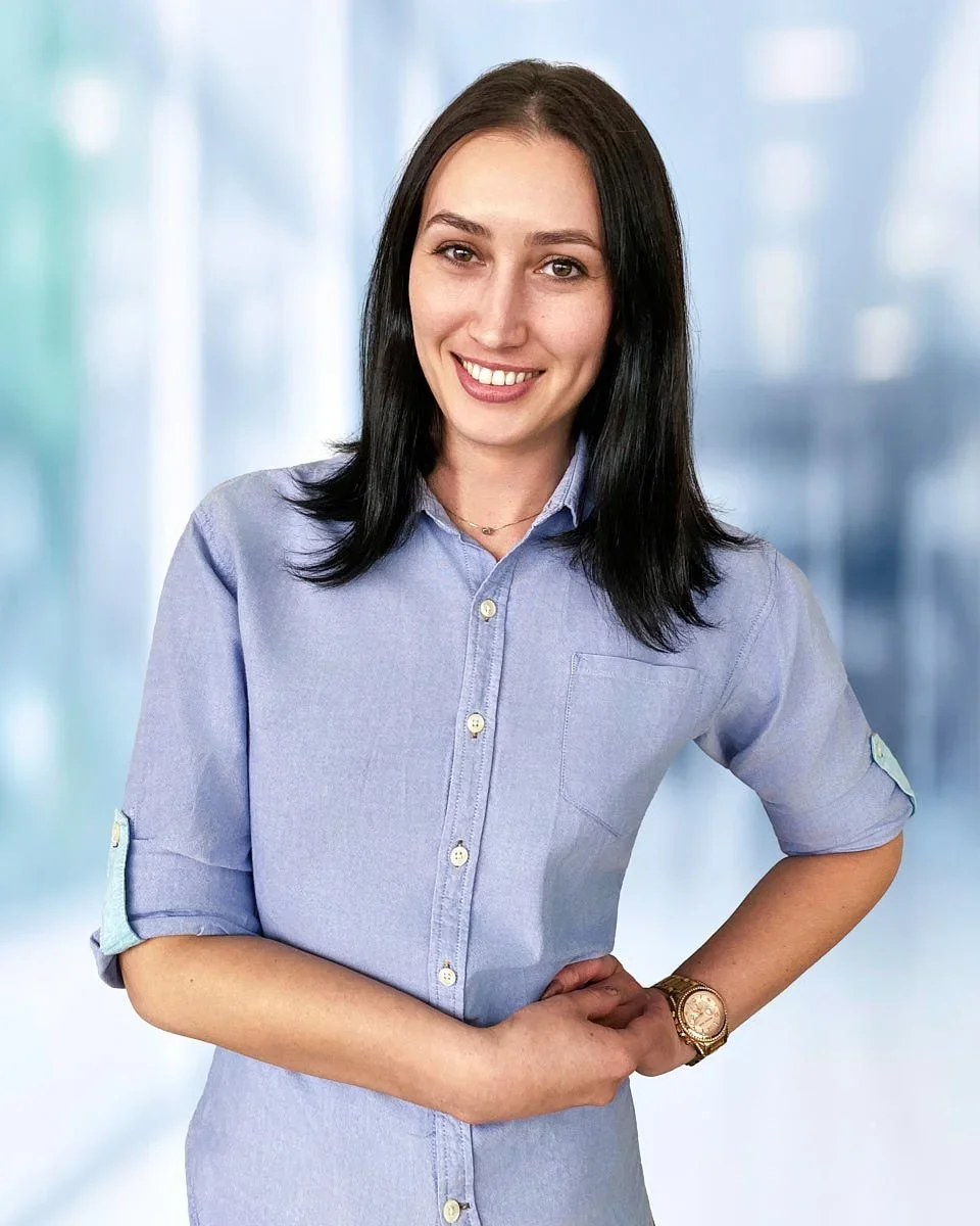A smiling woman with dark hair wearing a light blue button-up shirt standing in front of a blurred indoor background.