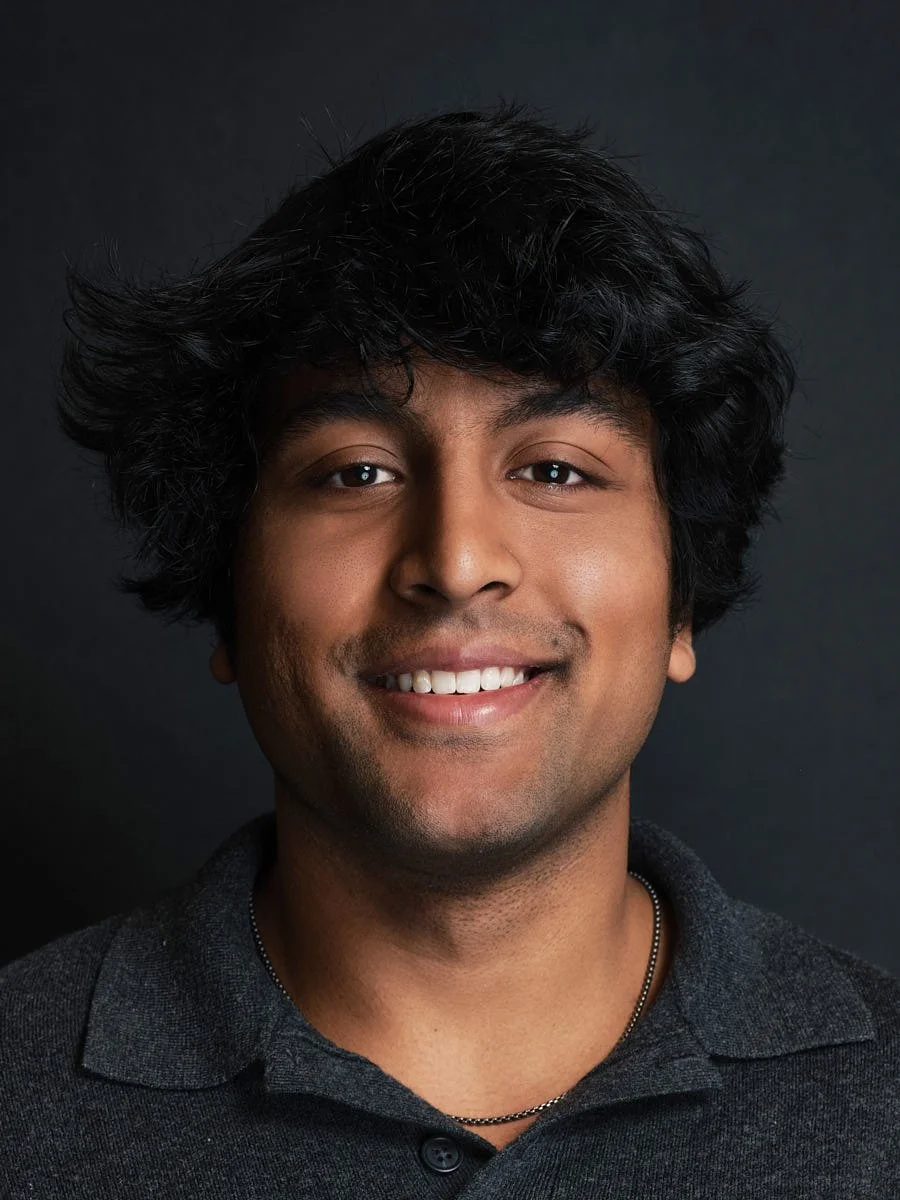 Headshot of a smiling man with dark, curly hair, wearing a gray collared shirt and a chain necklace against a dark background by Match Production.