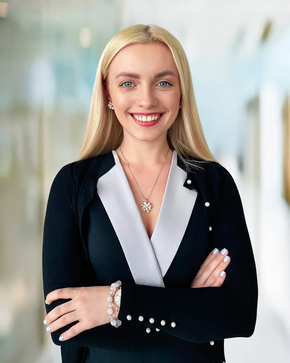 A woman with long blonde hair, blue eyes, wearing a black cardigan with pearl buttons, a white top, a pearl necklace, earrings, a watch, and a bracelet, smiling with her arms crossed in an indoor setting.