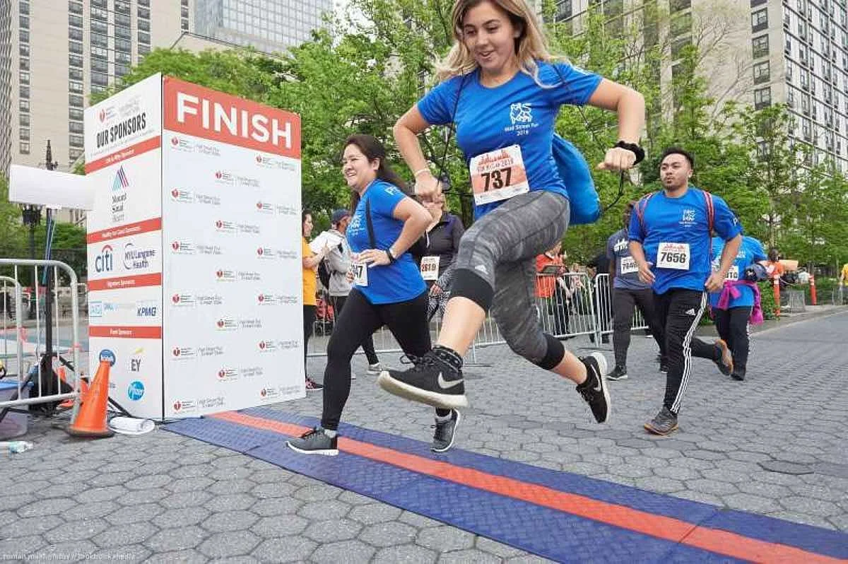 Two women and one man crossing the finish line at a race event in an urban park setting, with tall buildings and trees in the background. They are smiling and appear to be celebrating.