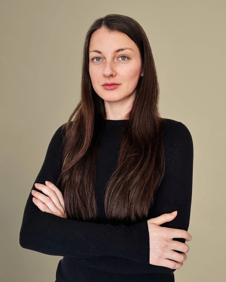 A woman with long brown hair, blue eyes, and fair skin standing with arms crossed, wearing a black long-sleeve shirt against a beige background.