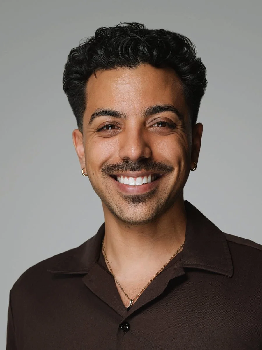 A smiling man with dark curly hair, tan skin, wearing a dark brown shirt, gold necklace, and earrings, posing against a gray background by Match Production.