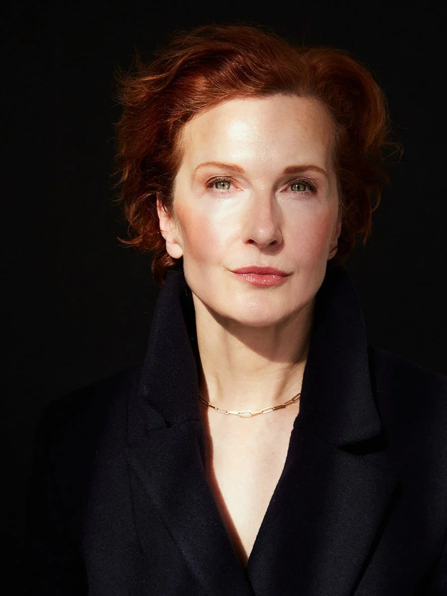 A woman with red hair and light skin wearing a black blazer and a small gold chain necklace, looking directly at the camera against a black background.