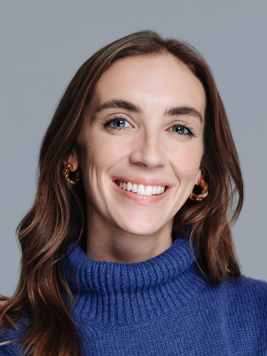 Close-up portrait of a smiling woman with blue eyes, brown hair, and gold earrings, wearing a blue turtleneck sweater, against a gray background by Match Production NYC.