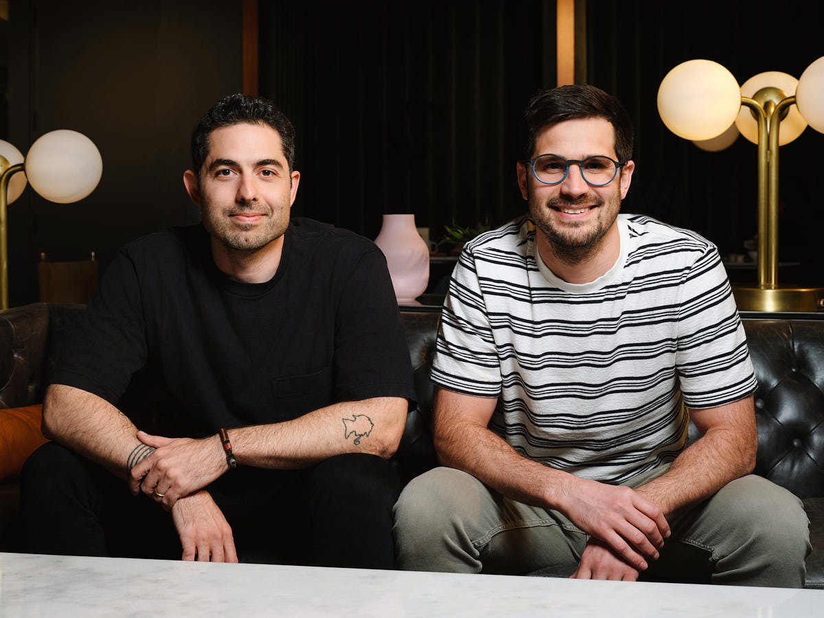 Two men are sitting on a black leather couch in a dimly lit room with modern decor. The man on the left has dark hair, a beard, and is wearing a black t-shirt. The man on the right has dark hair, glasses, a beard, and is wearing a striped t-shirt. Bo
