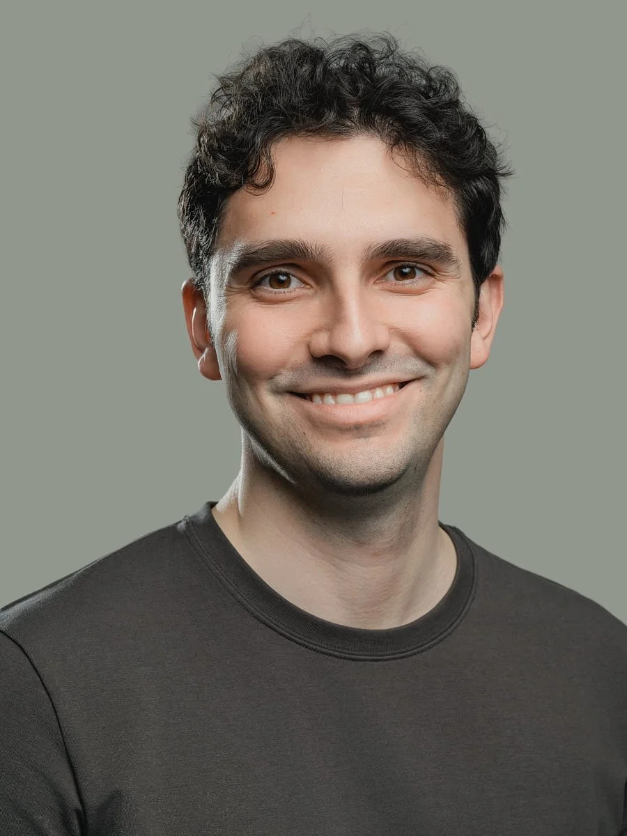 A smiling young man with dark, curly hair, wearing a black shirt, standing against a plain, light gray background by Match Production.