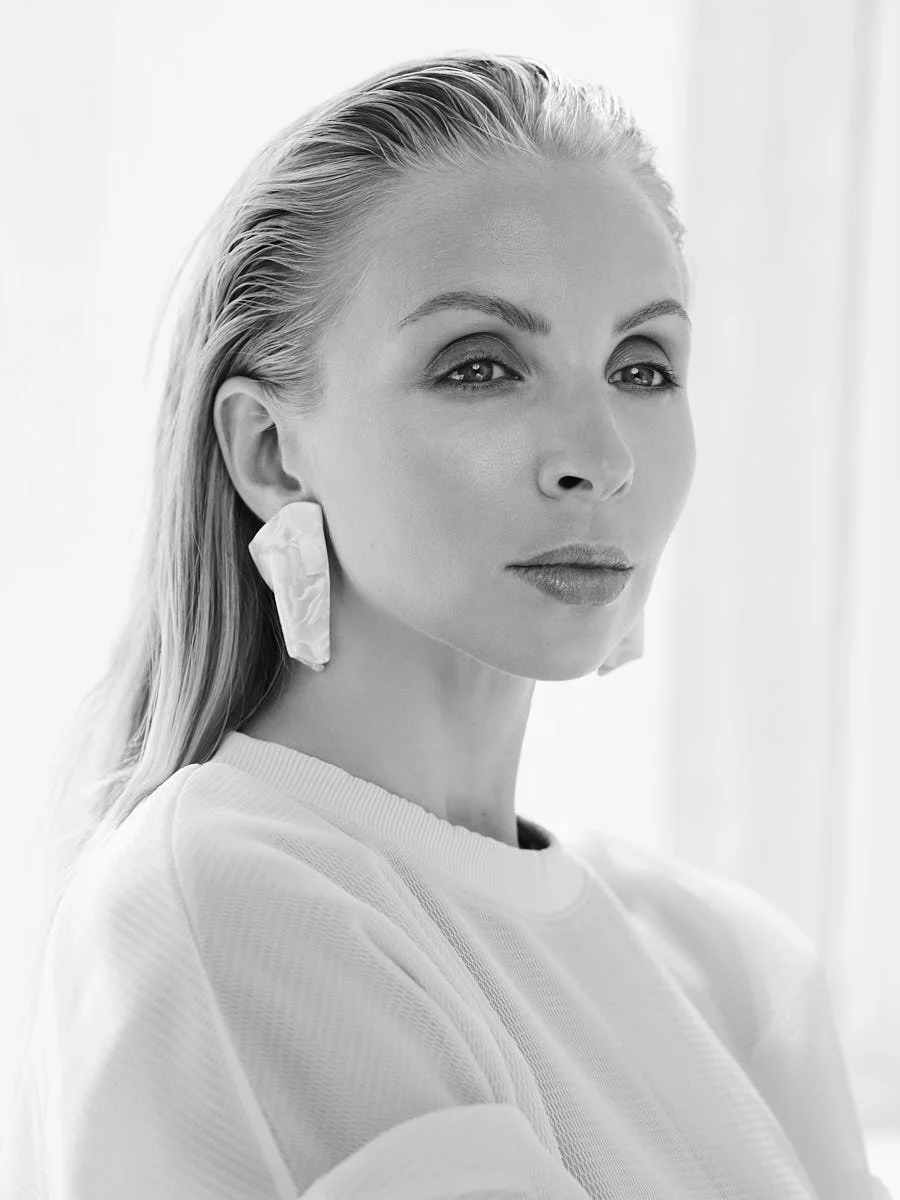 Black and white portrait of a woman with long blonde hair, wearing a white top and large earrings, looking slightly to the side with a serious expression.