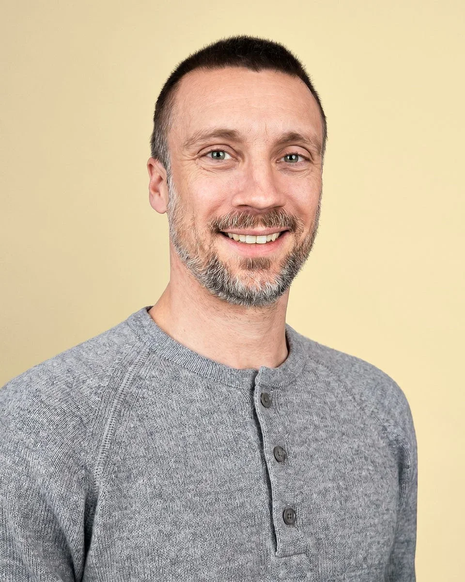 A smiling man with short dark hair, a beard, and blue eyes, wearing a grey button-up sweater, standing against a light yellow background.