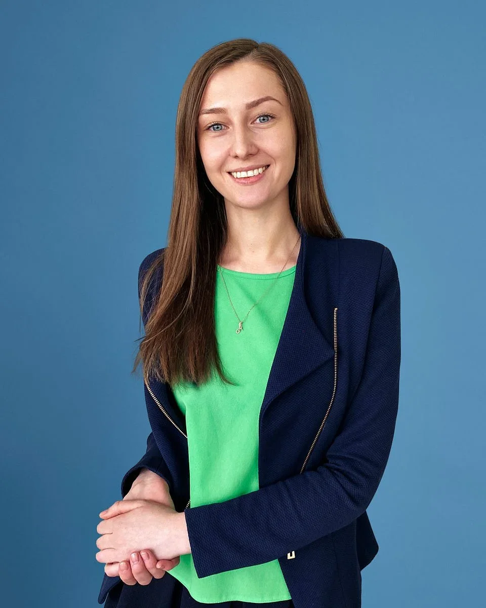 A young woman with long brown hair, blue eyes, and fair skin, smiling, standing against a blue background, wearing a green t-shirt, a navy blazer, and a silver necklace.