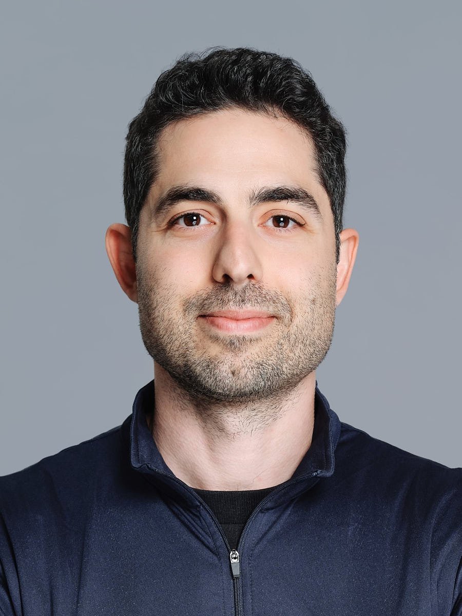 Headshot of a man with dark hair and light skin, wearing a navy blue zip-up jacket, smiling softly against a gray background by Match Production NYC.