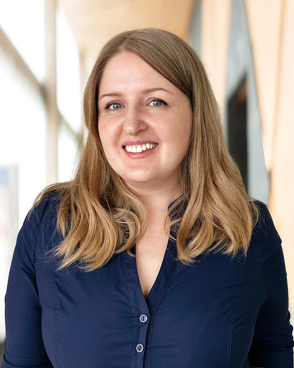 A woman with shoulder-length blonde hair, smiling, wearing a navy blue blouse, indoors with light-colored curtains and windows in the background.