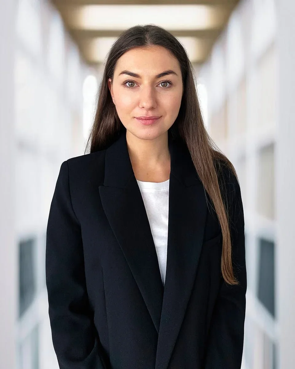 A young woman with long brown hair, wearing a black blazer over a white shirt, standing in a bright corridor with windows on both sides.