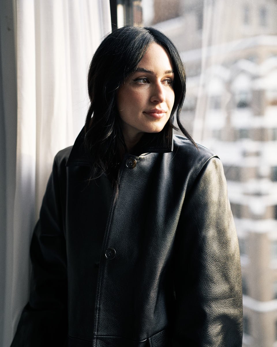 A young woman with black hair wearing a black leather jacket, looking out a window in a modern apartment, with city buildings visible outside by Match Production.