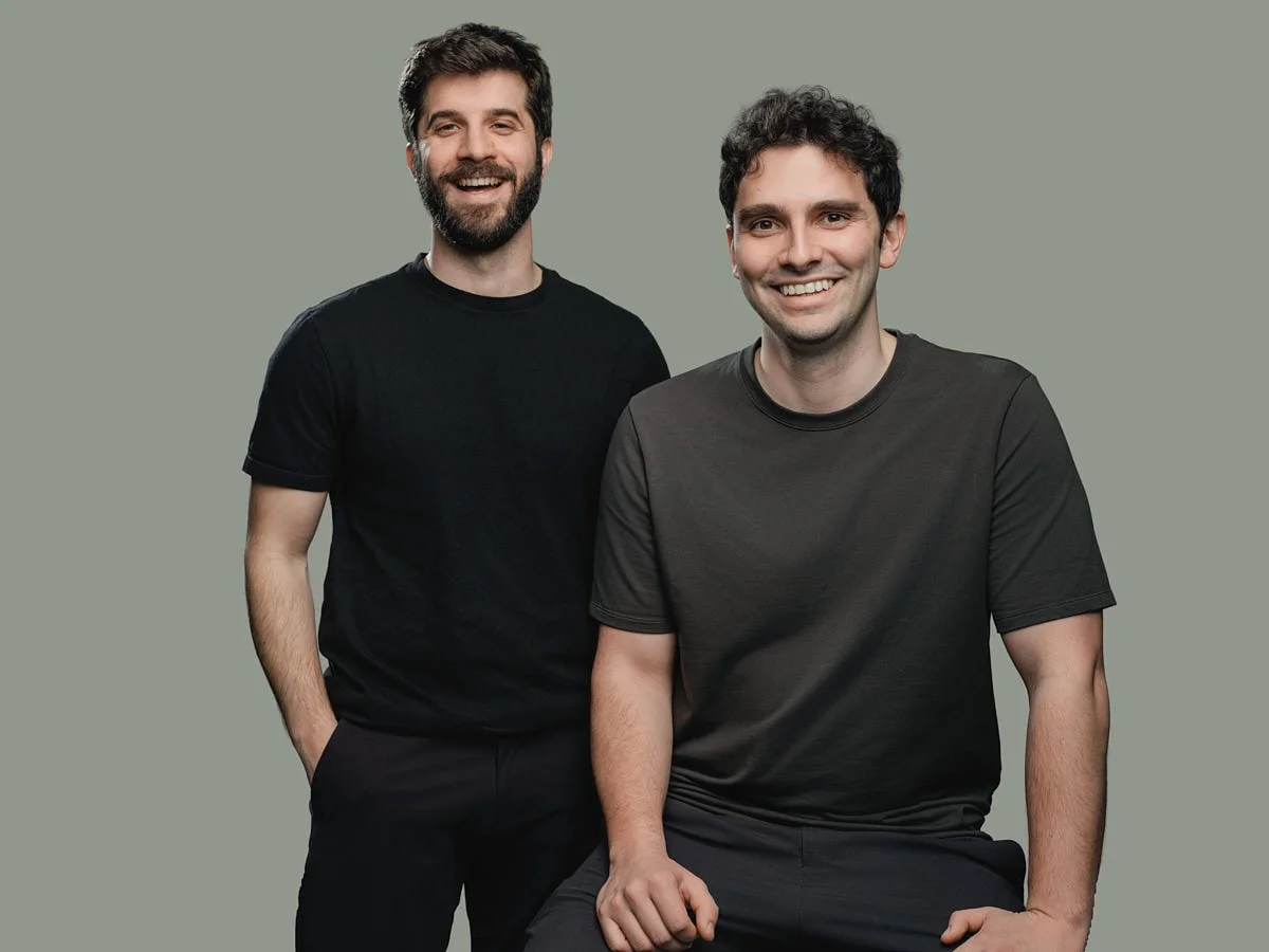 Two men smiling and posing against a plain gray background, one standing with his hand in his pocket and the other sitting with his hand resting on his knee by Match Production.