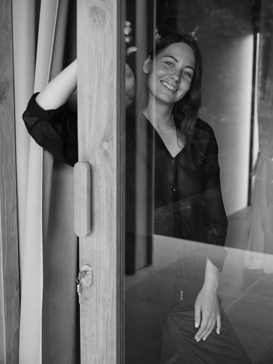 A woman with dark hair smiling, sitting inside, and seen through a glass window. She is wearing a dark blouse or dress, with her arm resting on a wooden surface. The photo is in black and white.