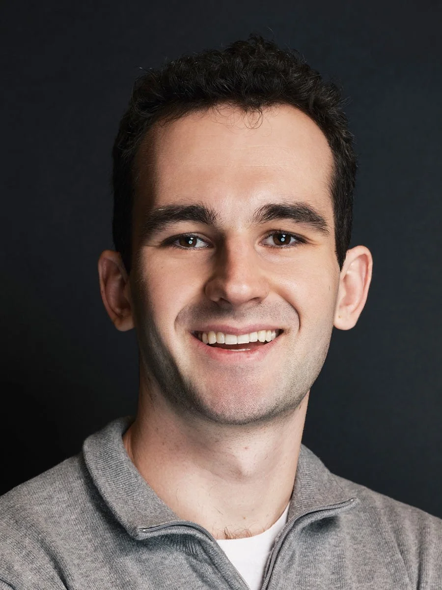 A young man with dark, curly hair, smiling, wearing a gray zip-up sweater against a dark background by Match Production.