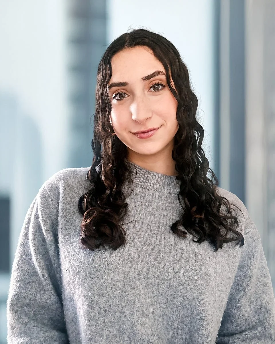 A young woman with curly dark hair and fair skin, wearing a gray sweater, standing indoors with a blurred background.