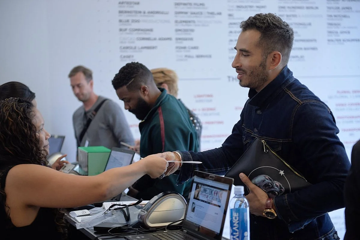 A man in a denim jacket accepting a ticket from a woman at a check-in counter, with several other people in line behind him in an airport or travel agency setting.