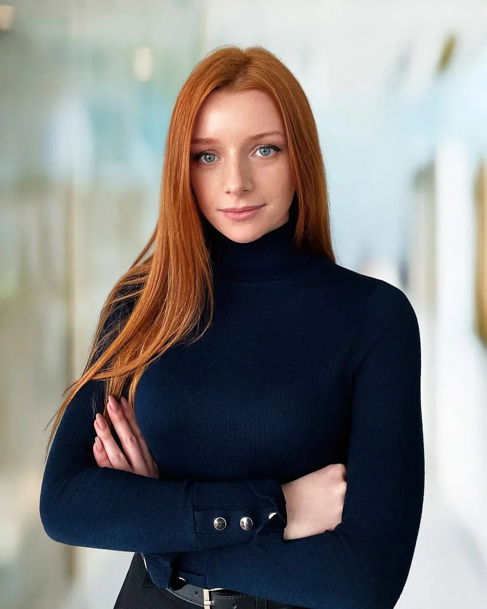 A woman with long red hair wearing a black turtleneck stands with arms crossed in a bright indoor setting.