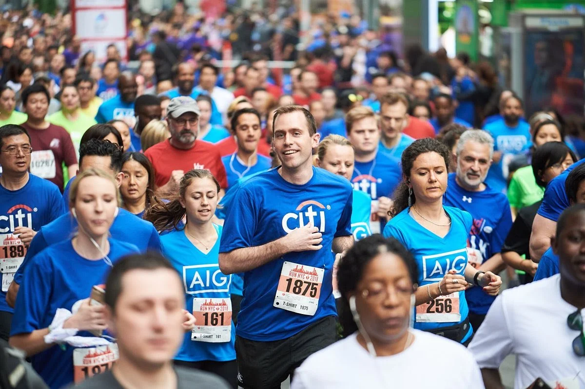 A large group of people participating in a marathon or running event, many wearing blue shirts with sponsorship logos, running together on the street.