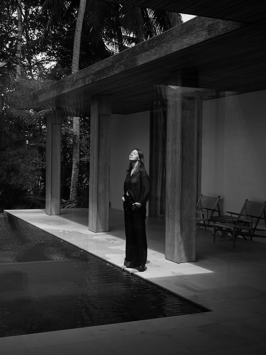 Woman standing on the edge of a swimming pool outside a modern house with wooden pillars and a covered patio, enjoying sunlight with her eyes closed.