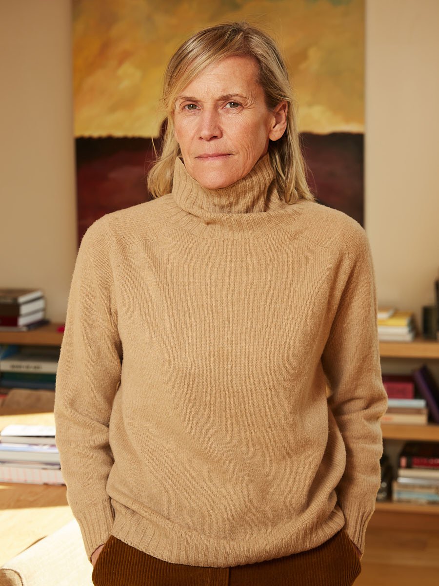 A woman with gray hair and a serious expression wearing a beige turtleneck sweater, standing in a room with bookshelves and a painting of a sunset or sunrise in the background.