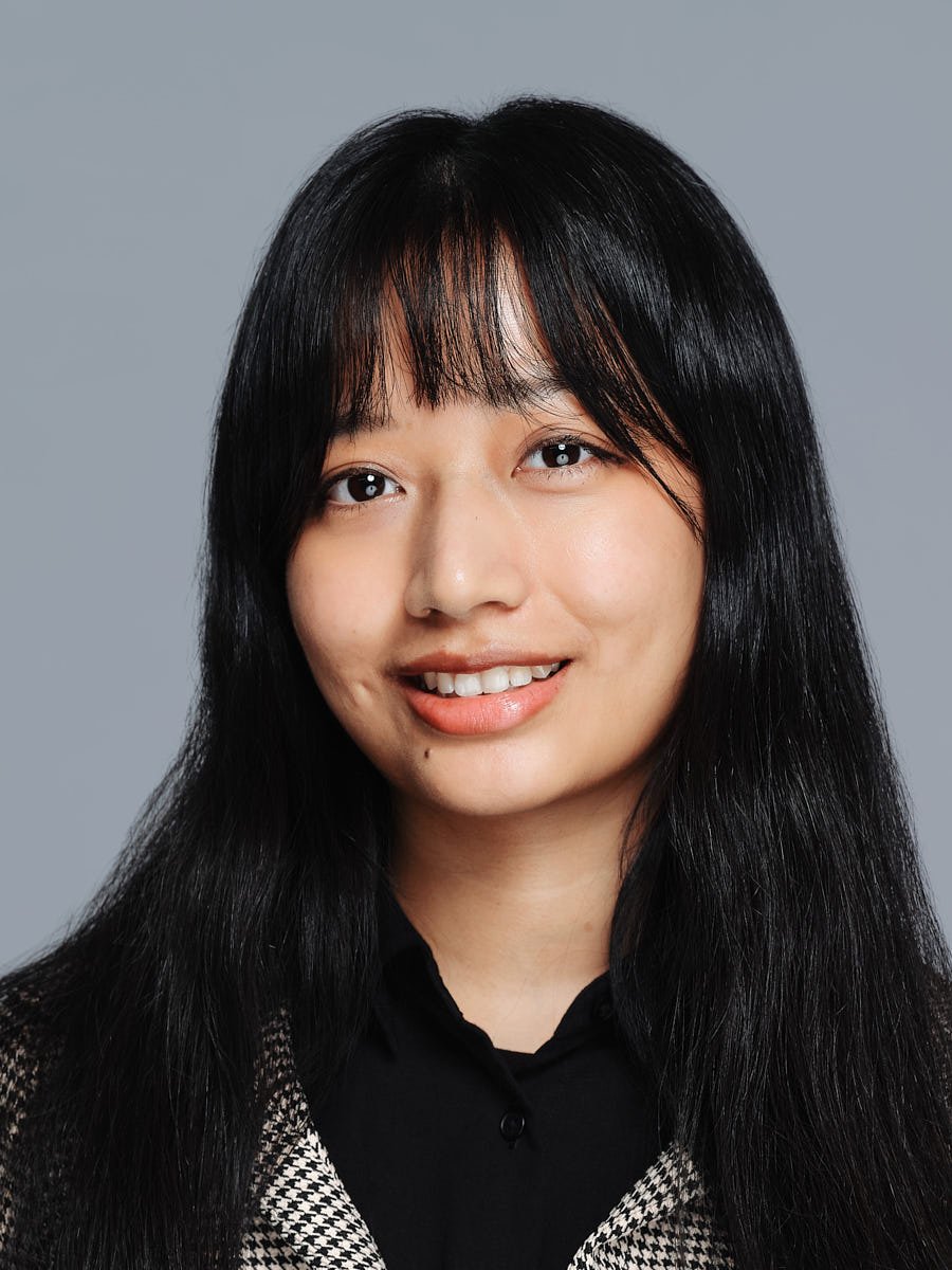 A portrait of an Asian woman with long black hair and bangs, smiling with teeth showing, wearing a black top and a black-and-white patterned jacket against a grey background by Match Production NYC.