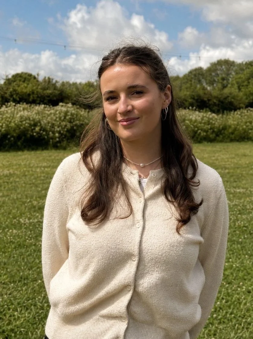 A smiling woman with dark brown hair in a bun, wearing a black blazer over a gray plaid dress, standing outdoors in a garden with green and purple plants behind her.
