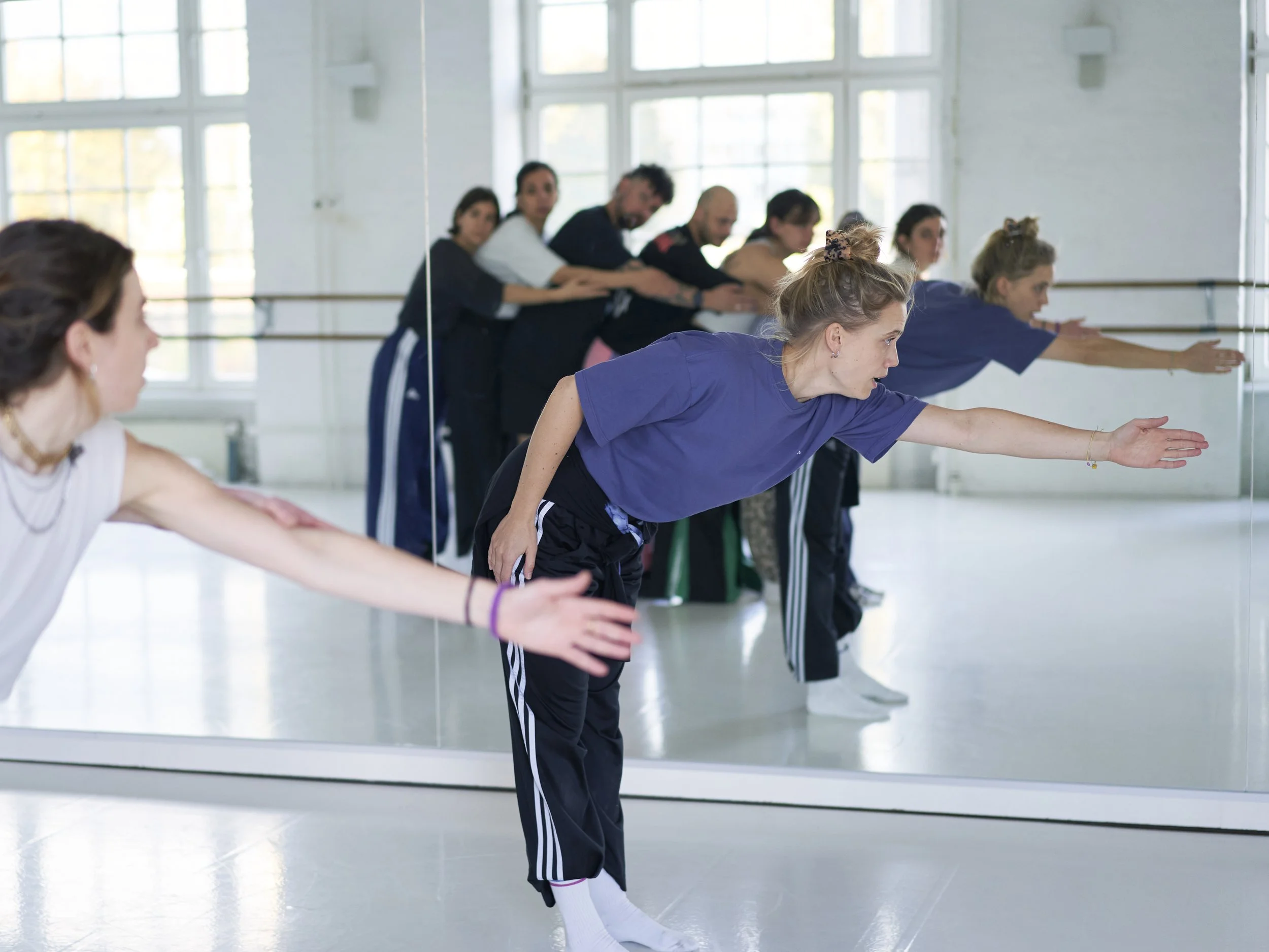Eine Gruppe von Menschen übt an der Ballettstange, darunter Frauen in sportlicher Kleidung und Männer, während sie auf einer spiegelbfeckten Tanzfläche in einem Studio mit großen Fenstern stehen.