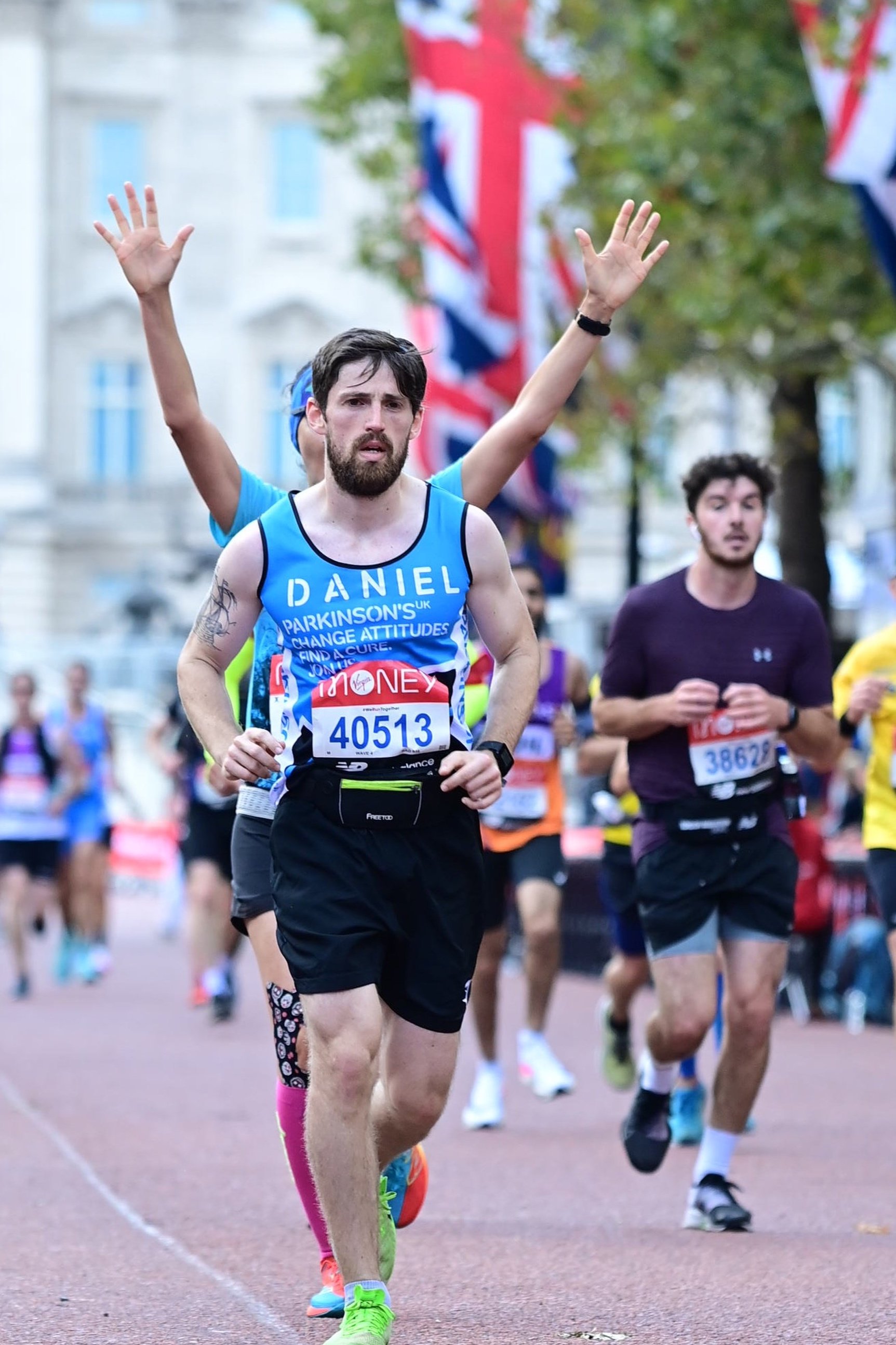 A man in a blue running shirt and black shorts running in a marathon, with his arms raised and a person behind him raising their arms in celebration, on a city street decorated with British flags.