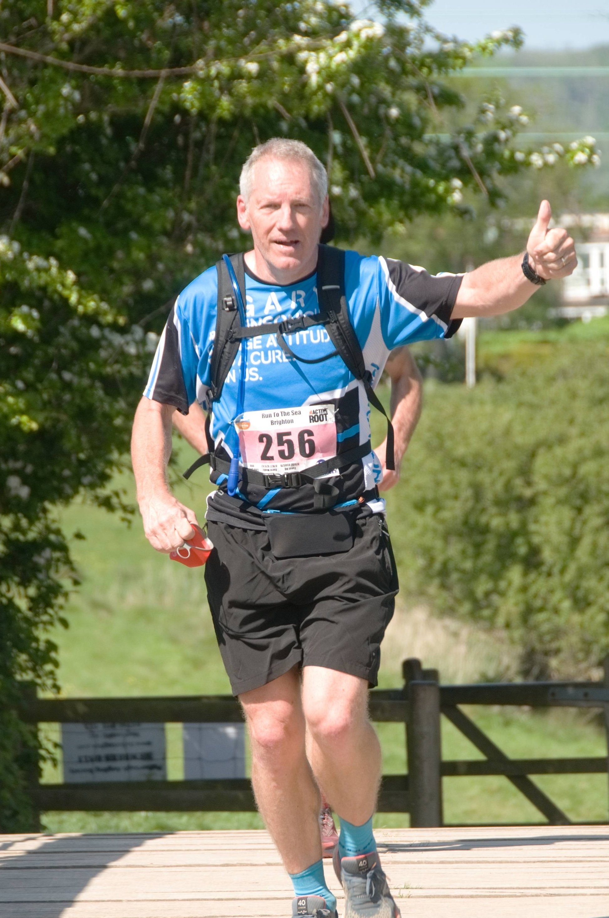 A middle-aged man running in a race, wearing a blue and black athletic shirt with race bib number 256, black shorts, and blue running socks, giving a thumbs-up while carrying a hydration pack, with green trees and a wooden fence in the background.