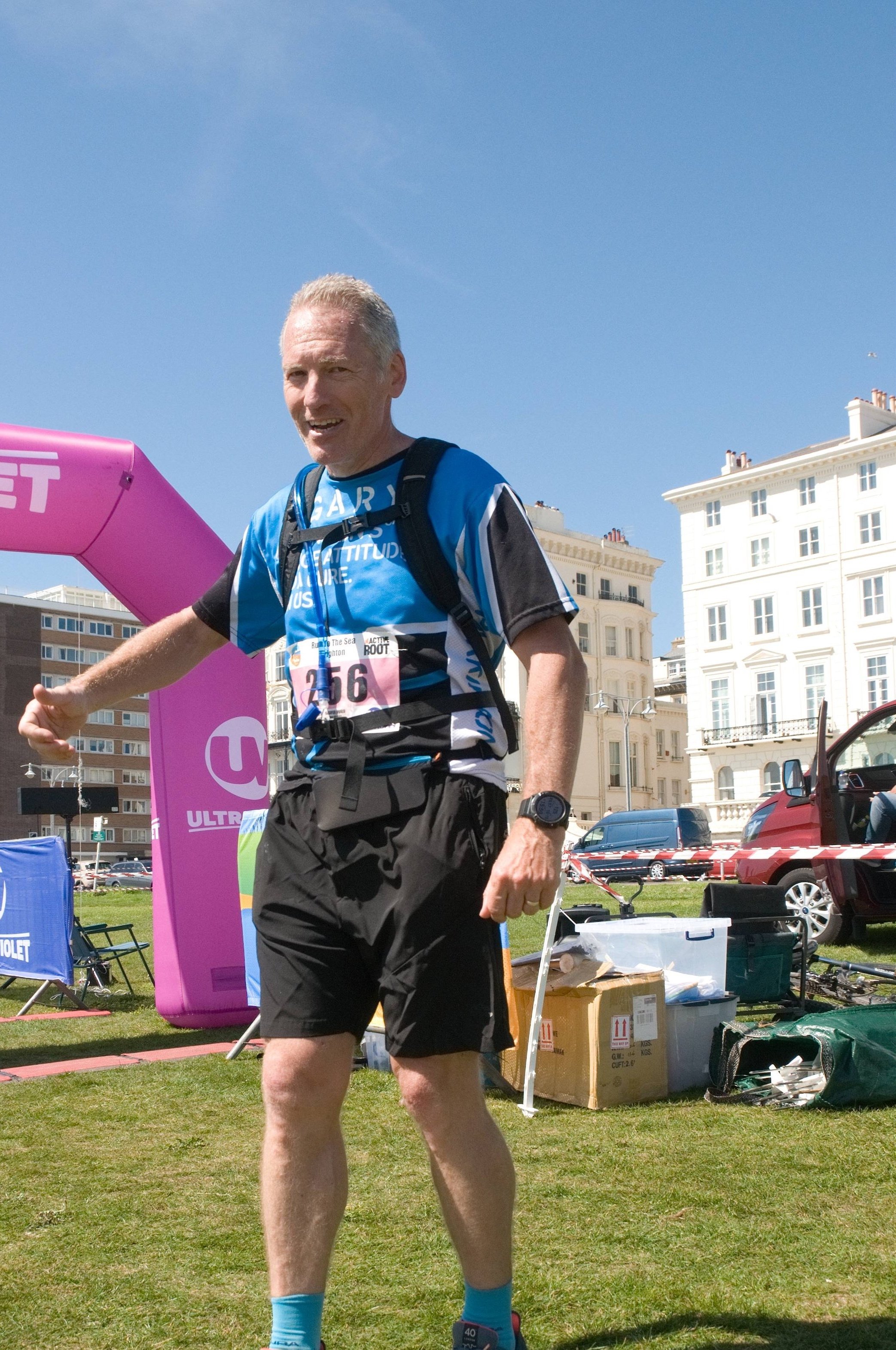 A man participating in a marathon, wearing running gear and a bib number 256, smiling, with a pink inflatable arch and buildings in the background on a sunny day.
