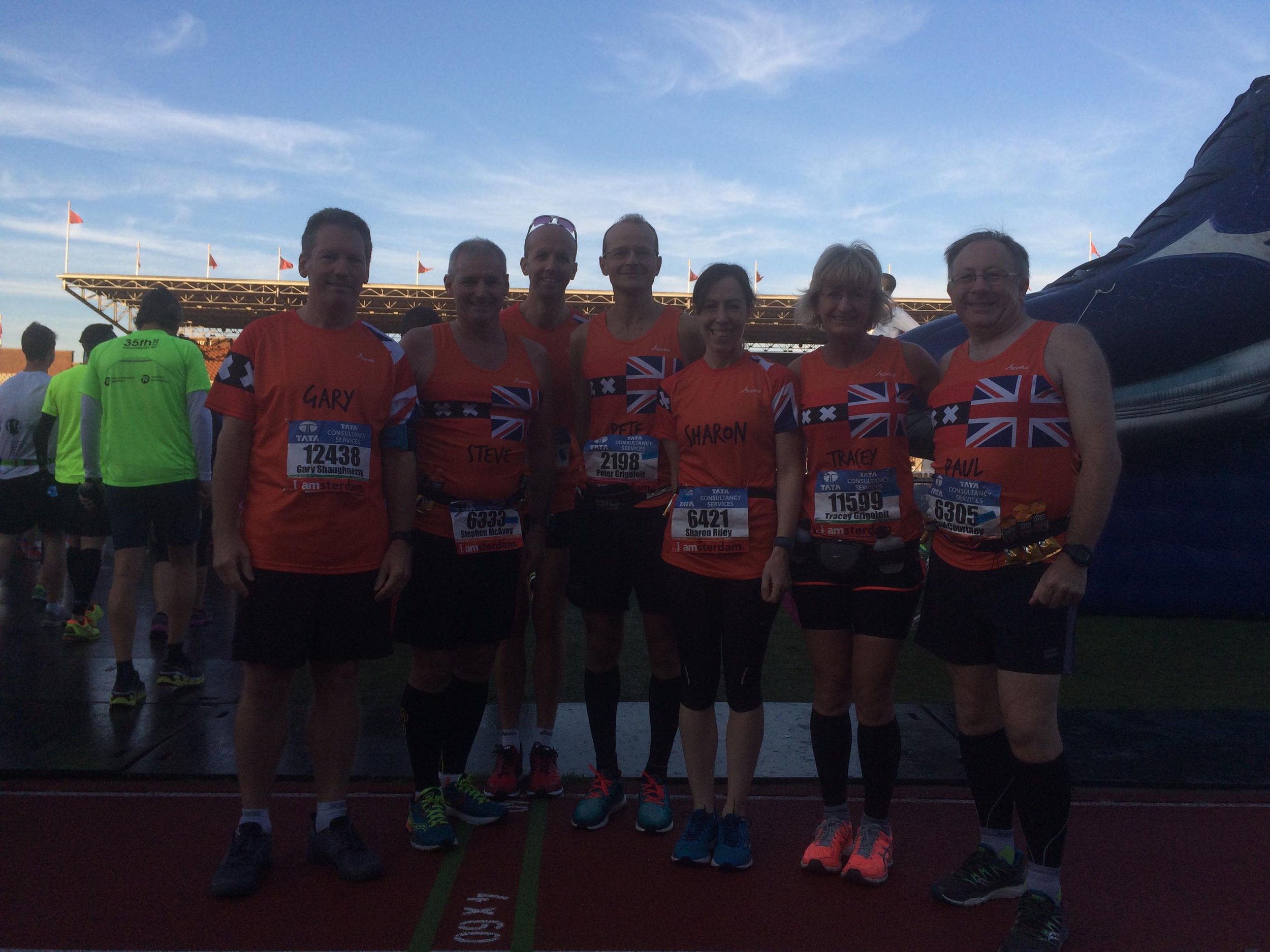 Group of marathon runners with race bibs, wearing matching orange shirts with UK flags, standing on a track at sunset.