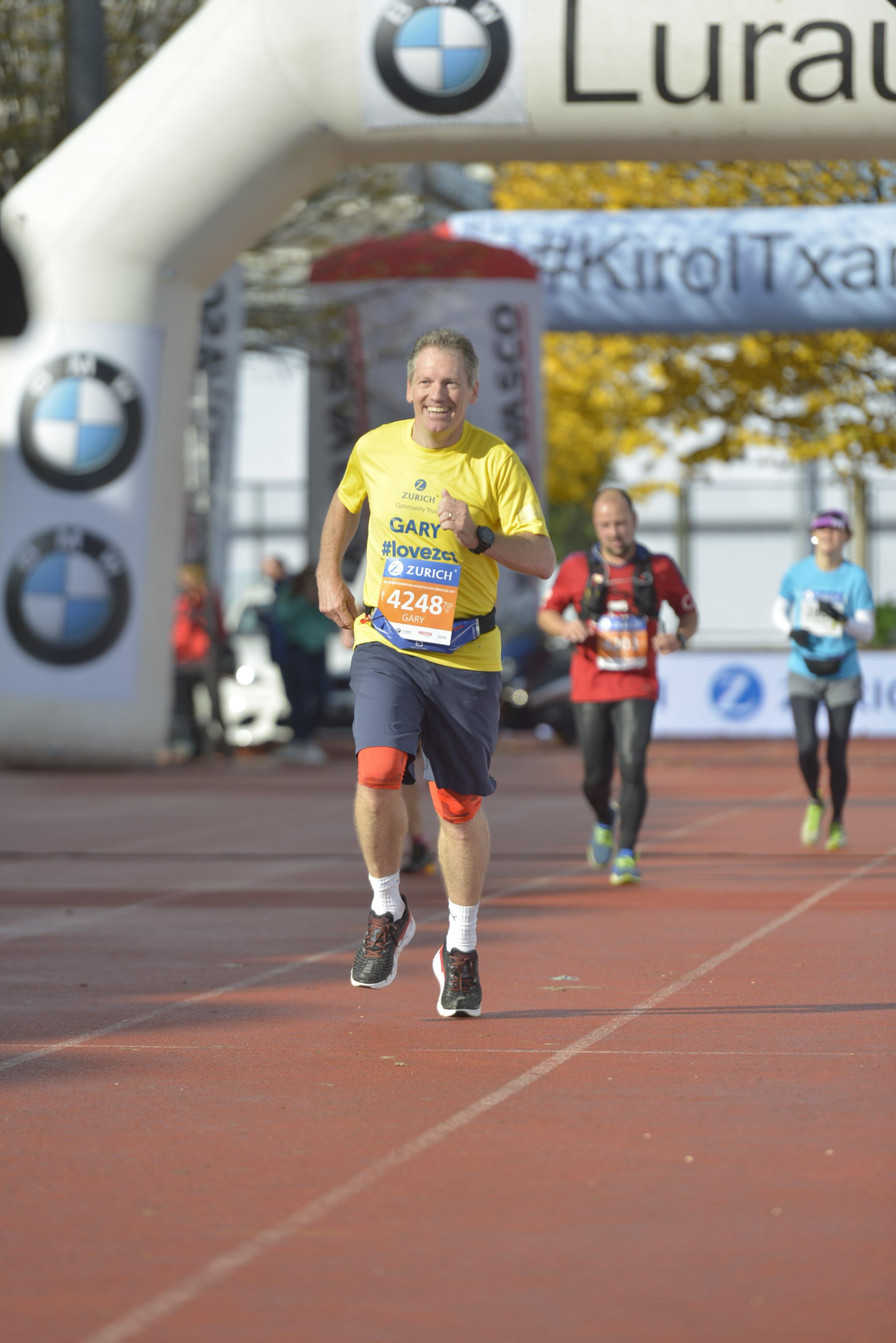 A man wearing a yellow shirt and black shorts running in a marathon, smiling as he approaches the finish line, with other runners behind him.