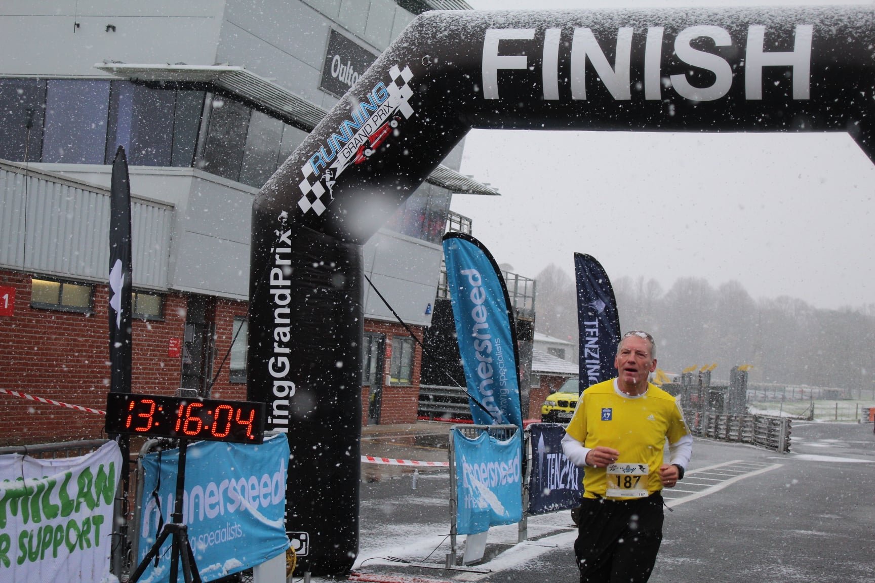 Male runner in yellow shirt crossing the finish line in snowy weather during a race. An inflatable arch with the word 'FINISH' and banners with race sponsors surround him. Snow is falling, and a digital clock shows the time 13:16:04.