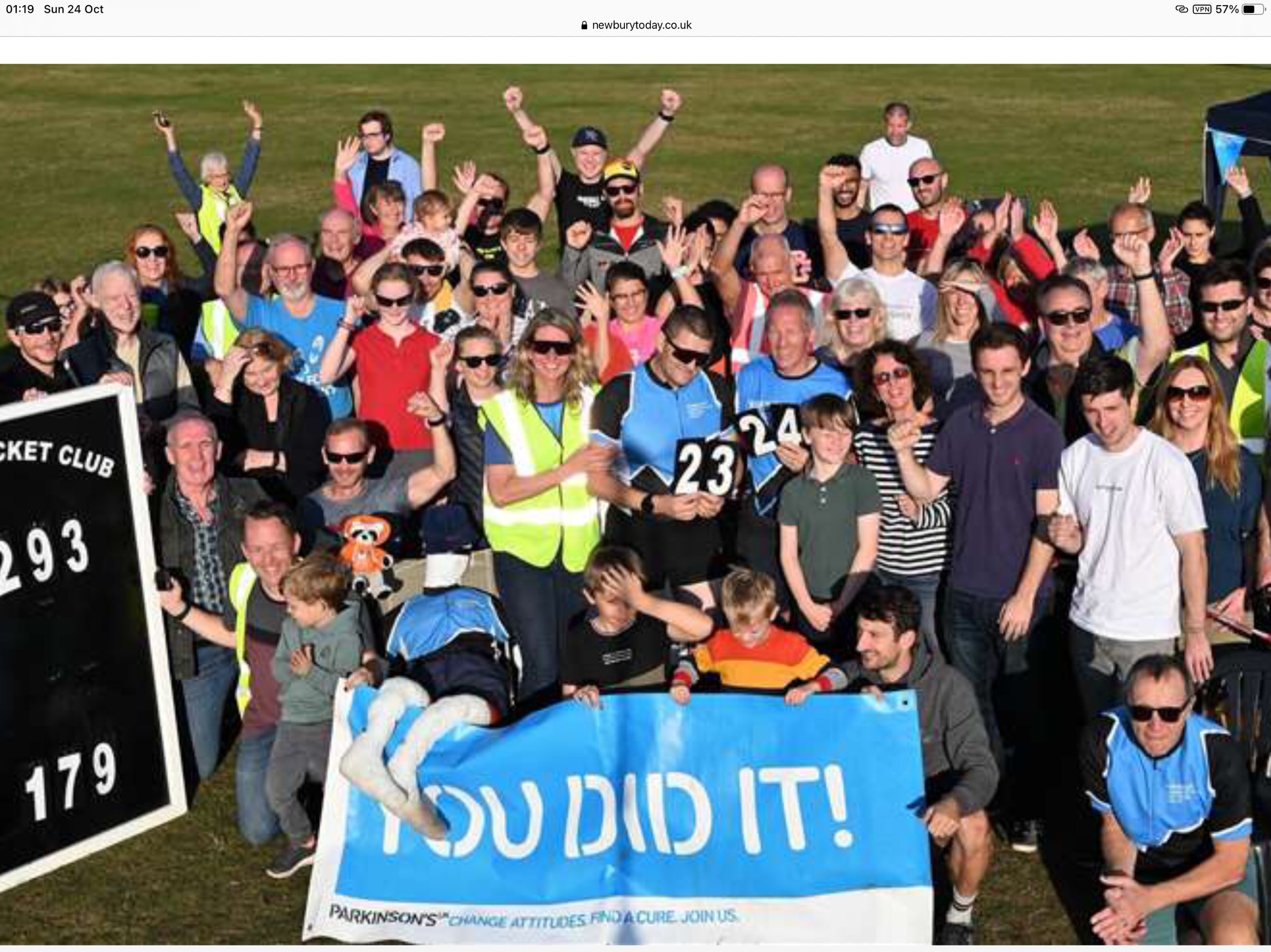 Group of people gathered outdoors during daylight, holding signs and banners, some wearing reflective vests, raising their hands, and smiling at the camera. There is a large blue banner with the message "You Did It!" and a smaller sign with numbers.