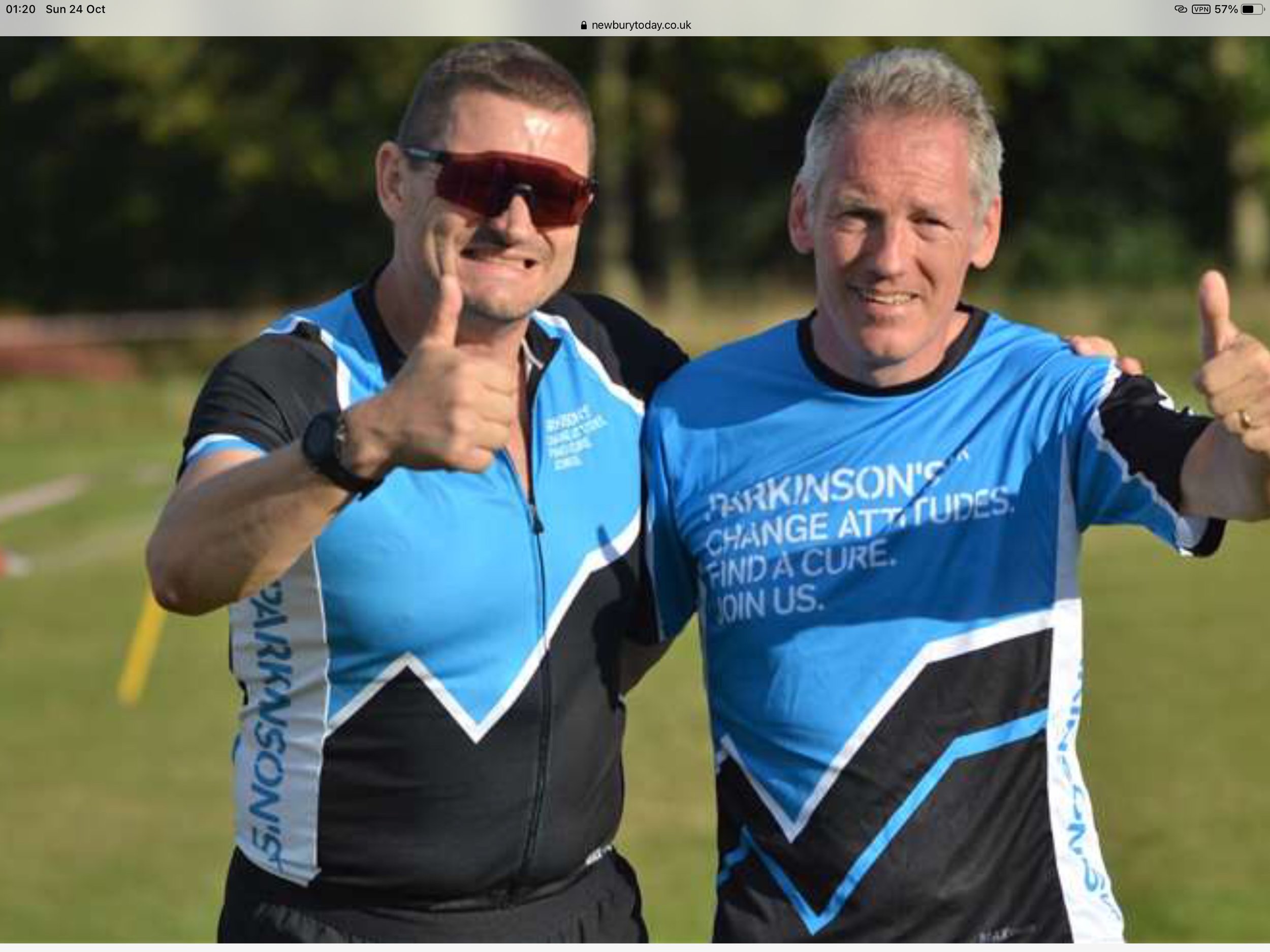Two men in blue sports shirts smiling and giving thumbs up, one wearing sunglasses, outdoors on a sunny day.