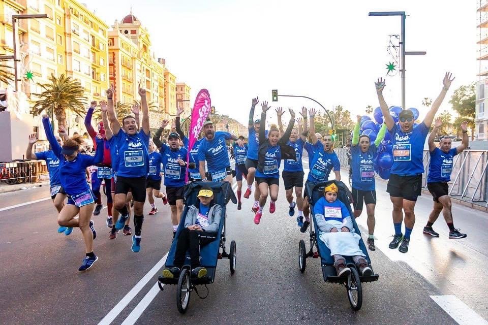 Group of runners participating in a marathon, some with children in strollers, all wearing blue running shirts, on a city street lined with buildings and palm trees.