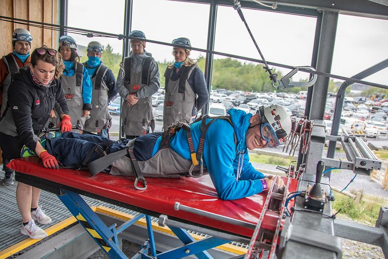A person in safety gear receives assistance from a staff member before a bungee jump experience, with several onlookers observing in the background.