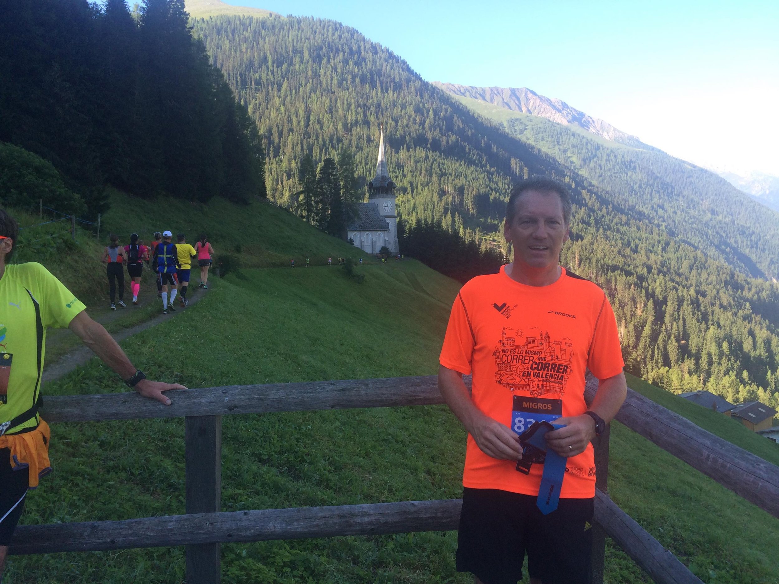Man in bright orange running shirt holding race bibs and medals, standing by a wooden fence in a mountainous area with a church steeple in the background, with other runners ahead on a trail.