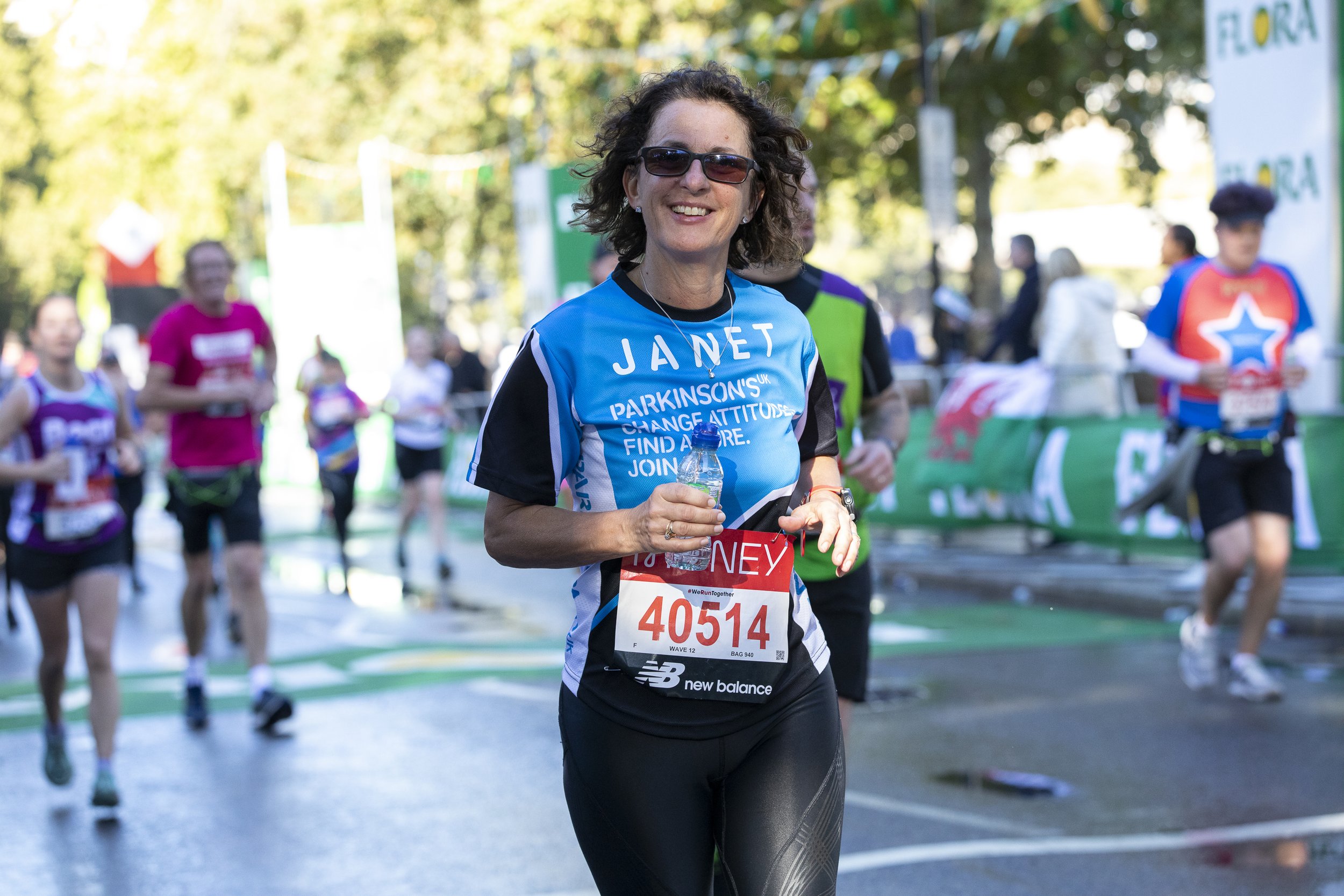 A woman running in a marathon, wearing a light blue race shirt with the name Janet on it, followed by other runners, at an outdoor race with trees and race banners in the background.