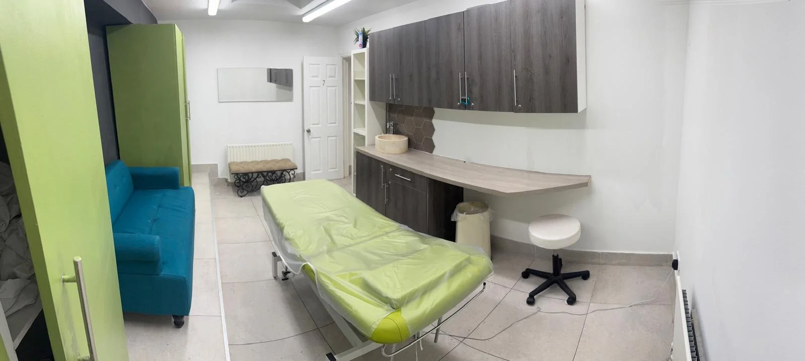 Medical examination room with a green examination table, a blue couch, cabinets, a mirror, a stool, and a sink.