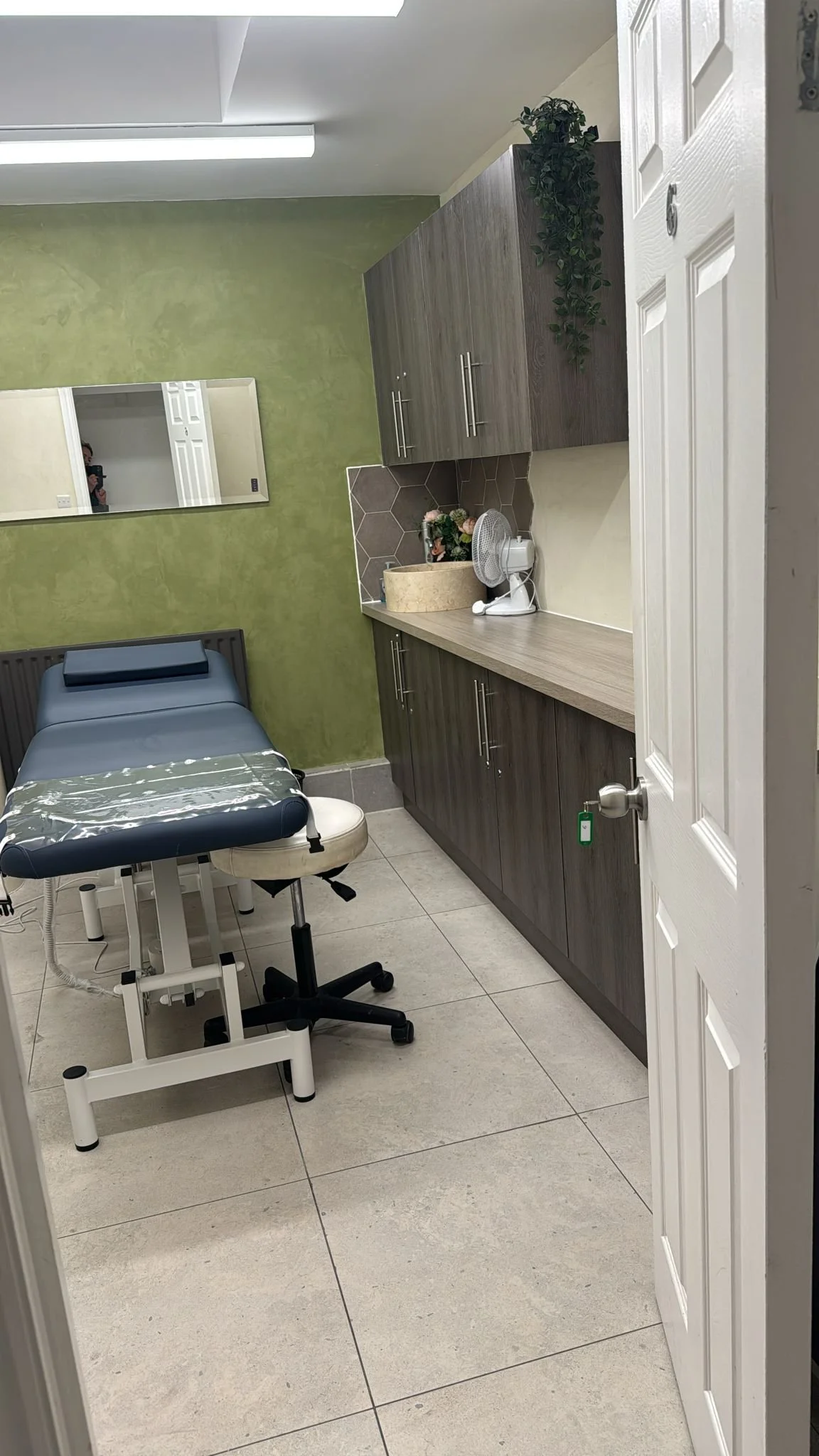 A medical examination room with a treatment table, a rolling stool, cabinets, a countertop with a fan, a plant, and a mirror on the green wall.